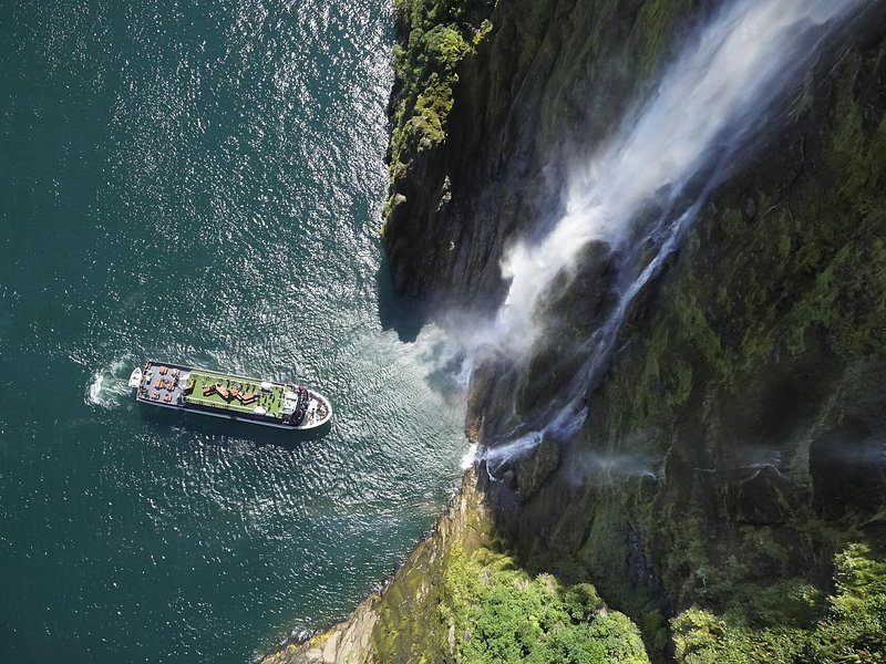 A cruise boat beside a waterfall falling into the fiord, seen from above