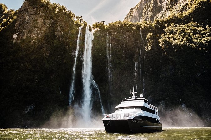 A dark-hulled boat nosing into the spray at the foot of a tall waterfall