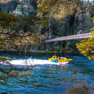 A yellow jet boat on a river below a swing bridge and beech forest