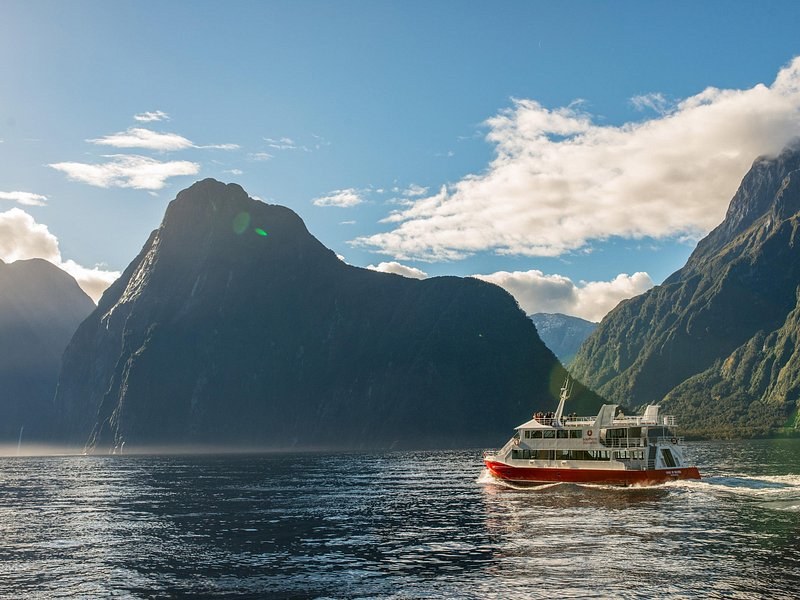 A red-hulled cruise boat heading out below a sheer dark peak and bright cloud