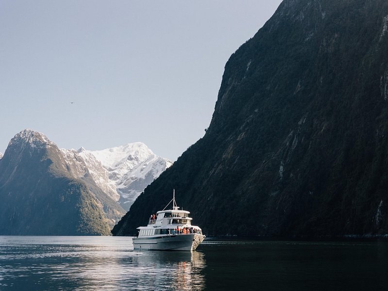 A small cruise boat below a sheer forested fiord wall, snow peaks behind