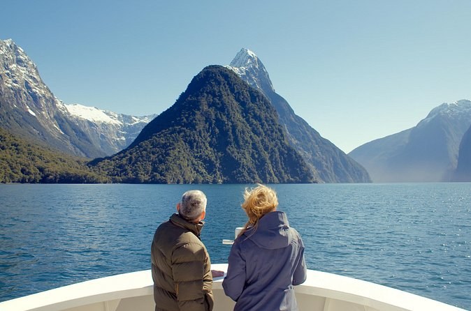 Two passengers at the bow of a cruise boat, facing a pointed peak across the fiord