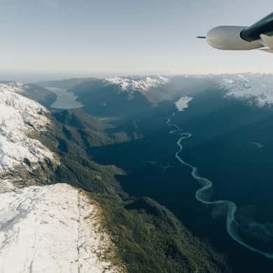 A braided river valley below snow slopes, seen past a plane's wing strut