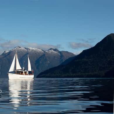 A two-masted yacht on a calm lake below forested hills